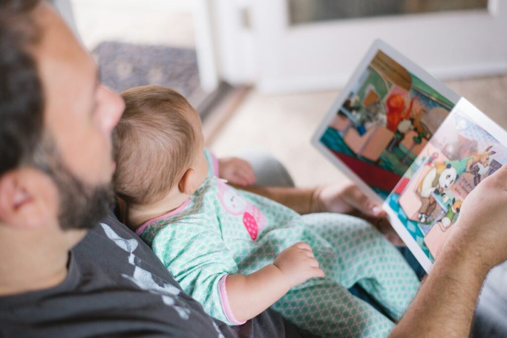 A child in pajamas reading a picture book with a parent on the edge of the bed